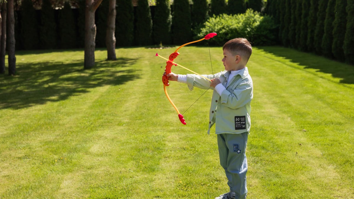 A young boy enjoying a sunny day playing with a toy bow and arrow on a grassy lawn.